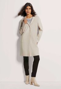 Beige strukturierter Mantel mit einem langen, geraden Design, zwei Seitentaschen und einem vorderen Verschluss. Kombiniert mit schwarzen Hosen und beigen Ankle-Boots.