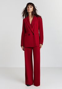 Roter, doppelt breiter Blazer mit schwarzen Knöpfen, aus glattem Stoff, sowie weite Hosen in passender Farbe, die einen maßgeschneiderten und kohärenten Look kreieren.