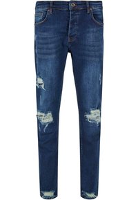 Jean en denim bleu avec une coupe slim, présentant des patchs usés et des bords effilochés. Comprend cinq poches et une fermeture à bouton.
