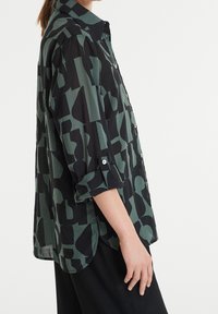 Femme portant une chemise ample à manches longues boutonnée avec des poignets retroussés dans un motif géométrique abstrait vert et noir.