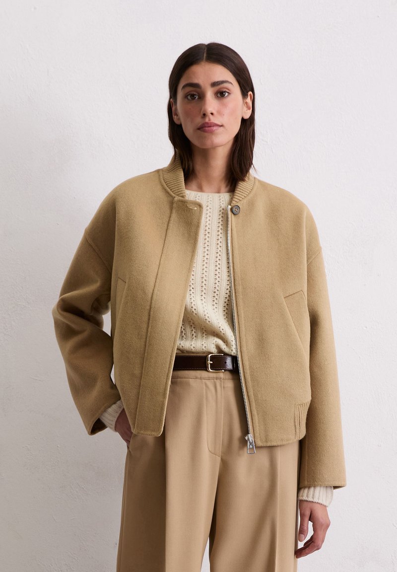 Tanfarbene Wollbomberjacke mit geripptem Kragen, Frontreißverschluss, Seitentaschen und lockerer Passform, kombiniert mit einem leichten Sweater und beigen Hosen.