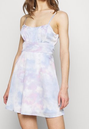 Frau in einem ärmellosen, pastellfarbenen Batik-Kleid mit dünnen Trägern, tailliertem Schnitt und ausgestelltem Rock vor einem schlichten Hintergrund.