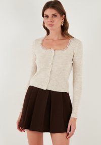 Ribota biskvitinė megztinė su banginiu apykaklės iškarpu ir mygtukų uždarymu. Suderinta su klostuota tamsiai ruda sijonu.