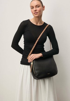 Nainen, jolla on musta pitkähihainen paita ja valkoinen hame, pitää mustaa nahkaista olkalaukkua, jossa on ruskea säädettävä olkahihna.