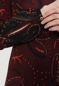 Robe bordeaux avec un motif paisley noir et orange, présentant un design à manches fendues et un accent de bouton décoratif au niveau du poignet.