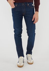 Jeans in denim blu scuro con vestibilità slim, caratterizzati da cuciture arancioni, cinque tasche e un'etichetta piccola alla vita. Indossati con scarpe da ginnastica bianche.