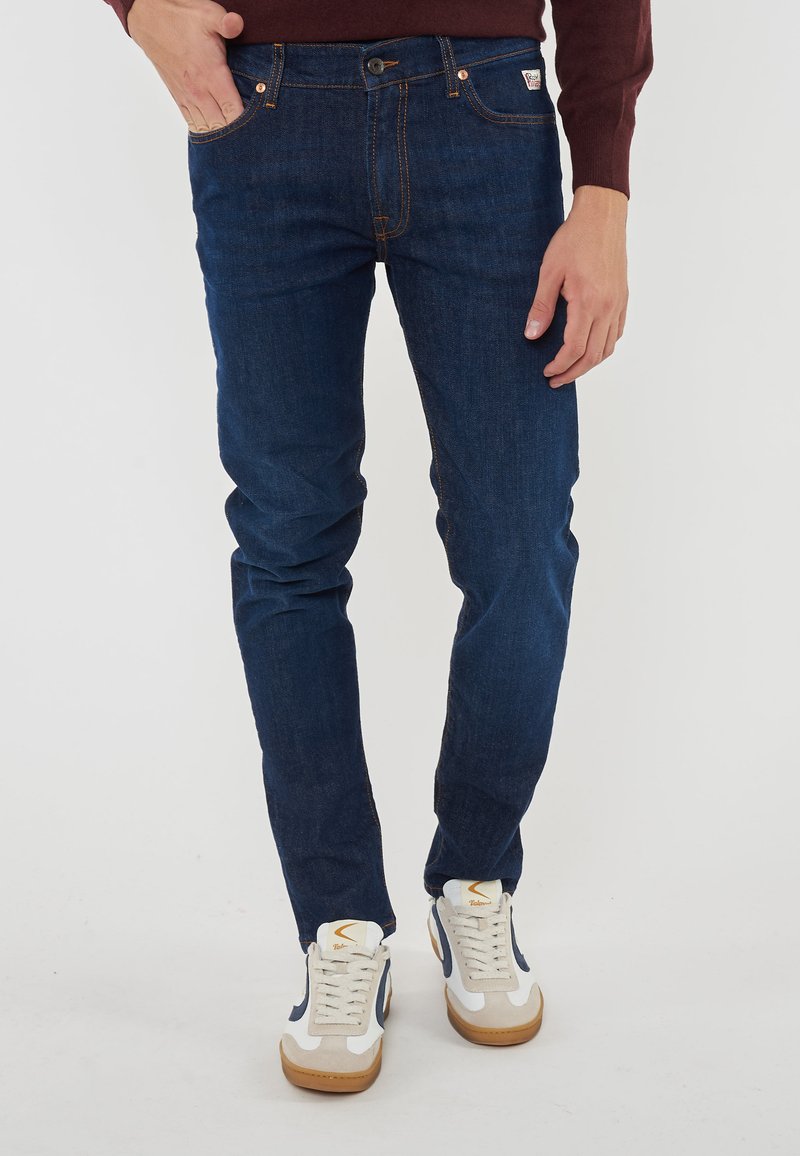 Jeans in denim blu scuro con vestibilità slim, caratterizzati da cuciture arancioni, cinque tasche e un'etichetta piccola alla vita. Indossati con scarpe da ginnastica bianche.