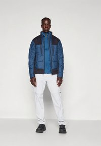 Mėlyna paminkštinta striukė su tamsiomis peties detalėmis, priekinių kišenių ir aukštu apykakliu; derinama su baltais kelnėmis ir juodais batais.