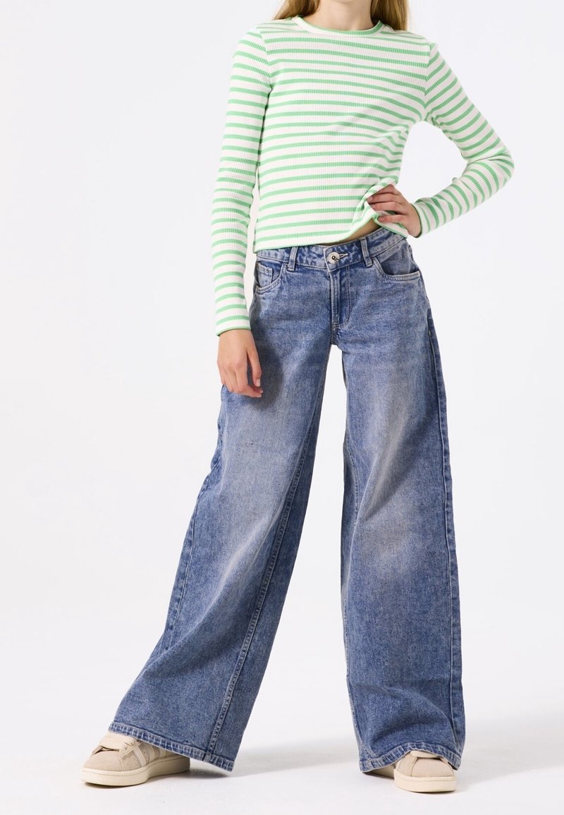 Un haut à rayures vertes et blanches à manches longues, associé à un jean en denim bleu à jambes larges. Le jean est doté d'une fermeture à bouton et de poches avant.