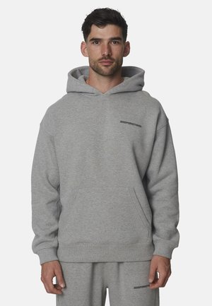 Uomo che indossa una felpa con cappuccio grigio chiaro e pantaloni della tuta abbinati, entrambi con un piccolo testo nero sul petto e sulla coscia.