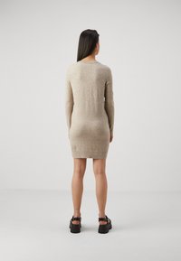 Vestito beige lavorato a maglia con maniche lunghe e scollatura rotonda, caratterizzato da una silhouette aderente e orlo a coste. Indossato con sandali neri con plateau.