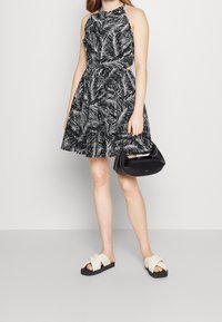 Černobílé květované šaty s halter výstřihem, staženým pasem a rozšířenou sukní, doplněné černou kabelkou a světlými sandály.
