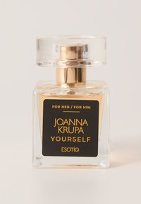 Przezroczysta szklana butelka perfum o kwadratowym kształcie, ze złotymi akcentami i czarną etykietą z napisem "JOANNA KRUPA YOURSELF ESOTIQ."
