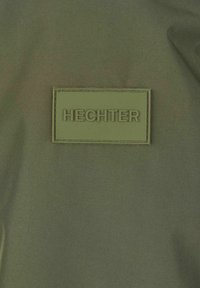 Grüner Stoff mit einer glatten Oberfläche zeigt ein rechteckiges Lederpatch, das mit "HECHTER" in Großbuchstaben geprägt ist.