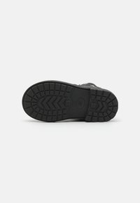 Semelle de chaussure en caoutchouc noir avec un motif de semelle texturé, présentant des rainures et des crampons pour une adhérence optimale. Un logo circulaire est visible au centre.