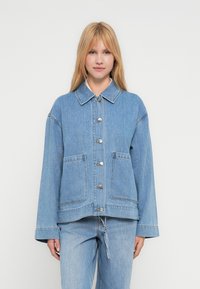 Kvinne iført lys blå denimjakke med frontlommer og sølvknapper, kombinert med matchende lys blå denimjeans.