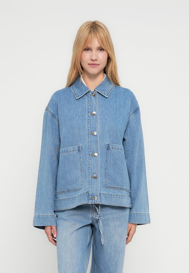 Kvinne iført lys blå denimjakke med frontlommer og sølvknapper, kombinert med matchende lys blå denimjeans.