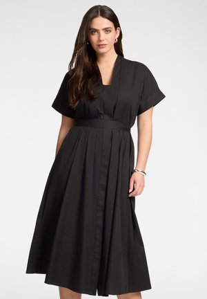 Kobieta o długich ciemnych włosach ubrana w czarną, plisowaną sukienkę midi z krótkim rękawem oraz srebrne kolczyki koła i bransoletki na jednolitym tle.