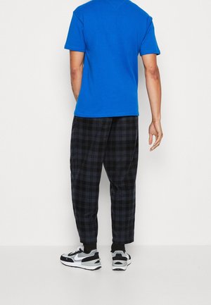 Homme portant une chemise à manches courtes bleue, un pantalon à carreaux noir et gris, des chaussettes noires et des baskets blanches et noires, debout contre un mur blanc.