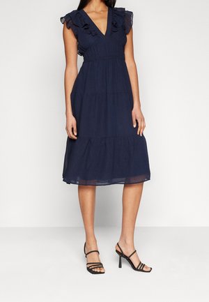 Kvinde iført en navyblå, knælængde kjole med flæser på ærmerne og sorte højhælede sandaler med flere remme.