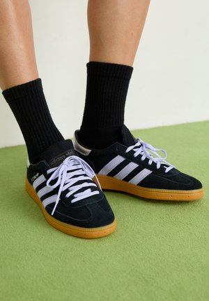 Nogi w czarnych butach Adidas z białymi paskami i gumowymi podeszwami, w połączeniu z czarnymi prążkowanymi skarpetami do kostek, stojące na zielonej podłodze.