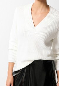 Valkoinen ribattu neulepaita, jossa on pitkät hihat ja V-kaula-aukko. Paita on rento ja siinä on taitetut hihan suut, yhdistettynä kiiltävään mustaan hameeseen.