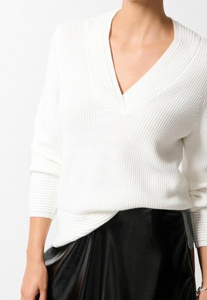 Pull en maille côtelée blanc à manches longues avec un col en V, présentant une coupe décontractée et des poignets repliés, associé à une jupe noire brillante.