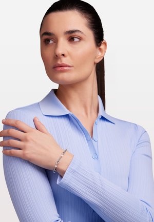 La maglietta a maniche lunghe di un blu chiaro a costine presenta un collo a camicia e chiusura con bottoni, abbinata a un bracciale in argento con piccoli dettagli.