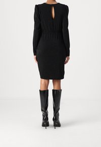 Robe noire à manches longues avec une silhouette ajustée, un tissu texturé et une ouverture en forme de clé à l'arrière, associée à des bottes brillantes arrivant jusqu'aux genoux.