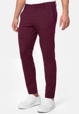 Pantaloni slim-fit bordeaux indossati da una persona in piedi abbinati a sneaker bianche basse e una camicia bianca.