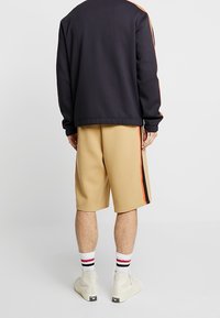 Hombre con una camiseta de manga larga negra y pantalones cortos beige hasta la rodilla con rayas laterales rojas y negras, calcetas blancas con rayas rojas y negras, y zapatillas blancas.