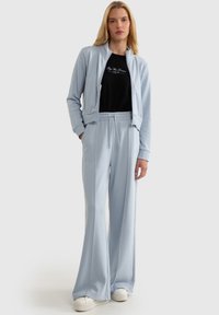 Gaiši zils sporta tērps, kas sastāv no zamšveida jaka ar rāvējslēdzēju un biksēm ar elastīgu jostu. Mīksts audums ar brīvu piegriezumu, kombinēts ar melnu t-kreklu.