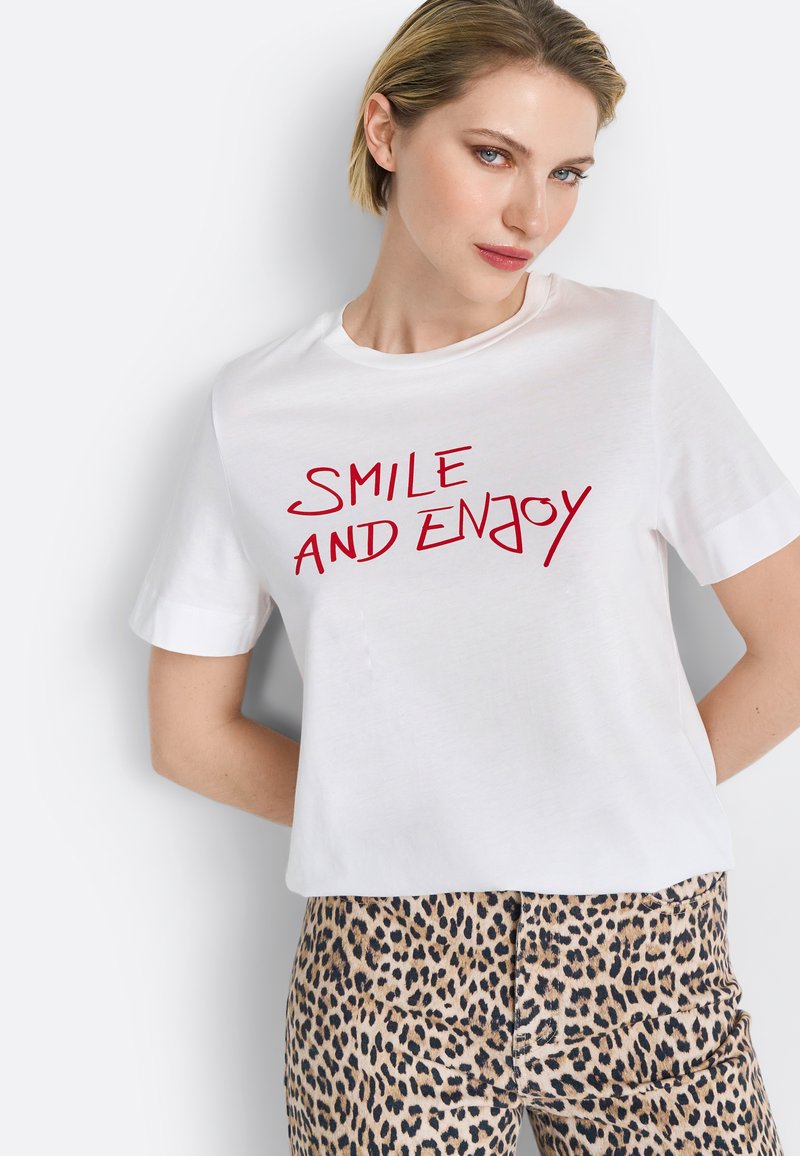 Witte katoenen t-shirt met een ronde hals en korte mouwen, met rode tekst die zegt "SMILE AND ENJOY." Gecombineerd met een broek met luipaardprint.