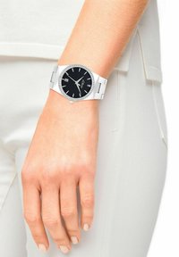 Silberne Analog-Uhr mit schwarzem Ziffernblatt, Kristallakzenten auf der Lünette, Metallband und einem Datumsdisplay um 6 Uhr.