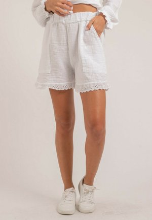 Femme portant un short blanc texturé avec des bords en dentelle festonnée, des poches et des baskets blanches, debout devant un fond uni.