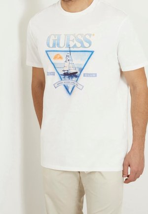 Man draagt een wit shirt met korte mouwen met een blauw driehoekig ontwerp waarop een zeilboot, zon en de tekst "GUESS YACHT CLUB" op de voorkant staan.