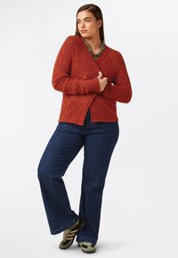 Rostfärgad stickad cardigan med ett strukturerat, chevronmönster, ihop med mörka denimflarebyxor och olivgröna sneakers.