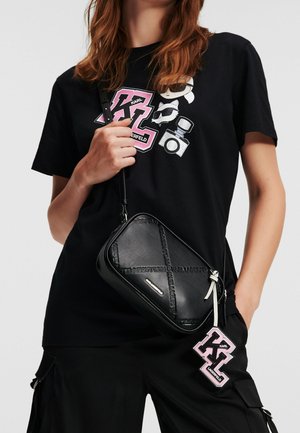Mujer con camiseta negra de Karl Lagerfeld con gráfico en rosa y blanco, bolso cruzado negro con el nombre de la marca en relieve y pantalones cargo negros.