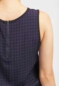 Tank top w kolorze fioletowym w wzór houndstooth, z czarnym wykończeniem i zamkiem błyskawicznym z tyłu. Materiał wydaje się gładki, o dopasowanym kroju i bez rękawów.