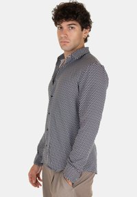 Giovane con i capelli ricci che indossa una camicia a maniche lunghe a fantasia geometrica bianca e nera e pantaloni beige, in piedi di lato.