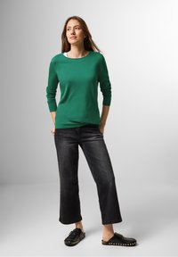 Grüner Strickpullover mit langen Ärmeln, kombiniert mit schwarzen weiten Jeans und schwarzen Slipschuhen mit goldenen Nieten. Schlichtes Design und lässige Passform.