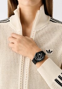 Montre noire avec un cadran rond et des marquages blancs sur un bracelet en tricot, portée sur une main au-dessus d'un sweat zippé crème avec des accents de rayures noires.