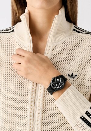 Montre noire avec un cadran rond et des marquages blancs sur un bracelet en tricot, portée sur une main au-dessus d'un sweat zippé crème avec des accents de rayures noires.