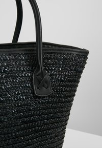 Sac cabas tissé noir avec texture tressée et poignées en cuir lisse cousues en forme de X sur un fond clair uni.