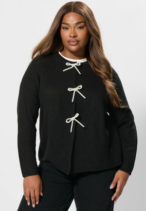 Sort strikket sweater med tre dekorative cremede sløjfer, rund halsudskæring, lange ærmer og en hi-low kant. Båret med mørke jeans.