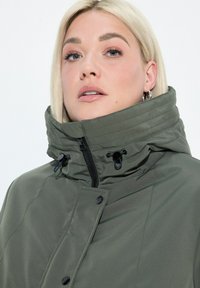 Blondynka w srebrnych kolczykach w kształcie kółek oraz oliwkowozielonej kurtce z wysokim kołnierzem, z detalami w postaci zamka i guzików, spoglądająca lekko do góry.