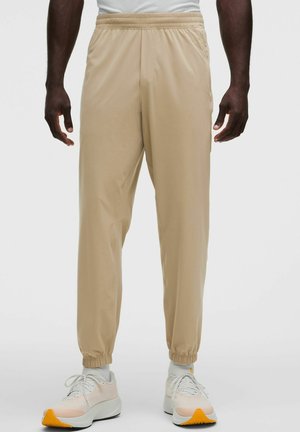 Uomo che indossa pantaloni da jogging beige con vita elastica e sneakers bianche con suole arancioni, in piedi su uno sfondo neutro.