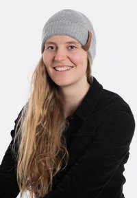 Szary dziany czapka z prążkowanym wzorem i brązową skórzaną metką. Model nosi czarną kurtkę z kordurowego materiału. Widoczne długie, falowane włosy.