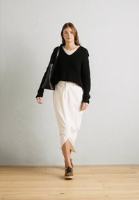 Svart strikket genser, kremfarget skjørt med en sideslitt, kombinert med brune sko. Modellen har en svart tote bag og går på et tregulv.