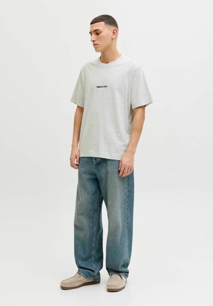 Graues Baumwoll-T-Shirt mit Rundhalsausschnitt und lockerem Sitz, mit schwarzem Text; kombiniert mit blauen weiten Jeans und beigen Slip-On-Schuhen.
